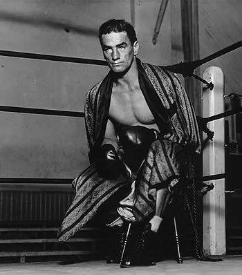 Frankie_Campbell_boxer_gospin Black-and-white photo of boxer Frankie Campbell seated in a boxing ring corner, wearing boxing gloves and a robe draped over his shoulders, looking toward the camera.